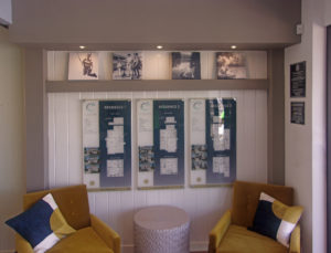 Interior wall with 2 seating chairs & end table. Behind them a display of 3 models on printed flat acrylic. Lifestyle photos displayed on bookshelf above.