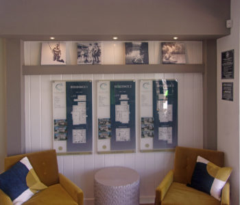 Interior wall with 2 seating chairs & end table. Behind them a display of 3 models on printed flat acrylic. Lifestyle photos displayed on bookshelf above.