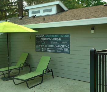 Pool capacity sign installed on wall, next to 2 lounge chairs & a umbrella.